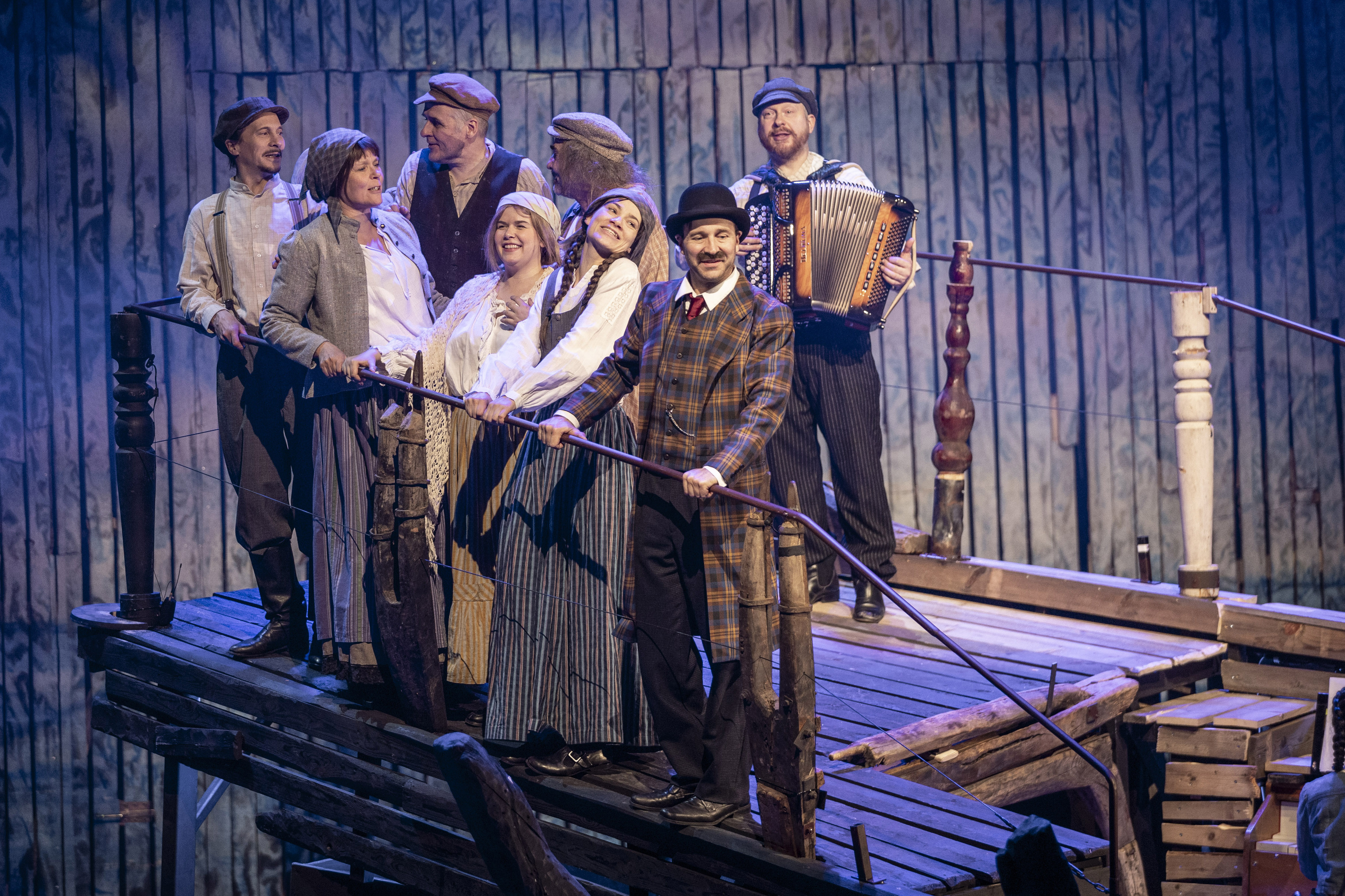 Kahdeksan näyttelijää, jotka ovat pukeutuneina 1900-luvun alun vaatteisiin seisovat laivankeulaa muistuttavassa lavasteessa. 